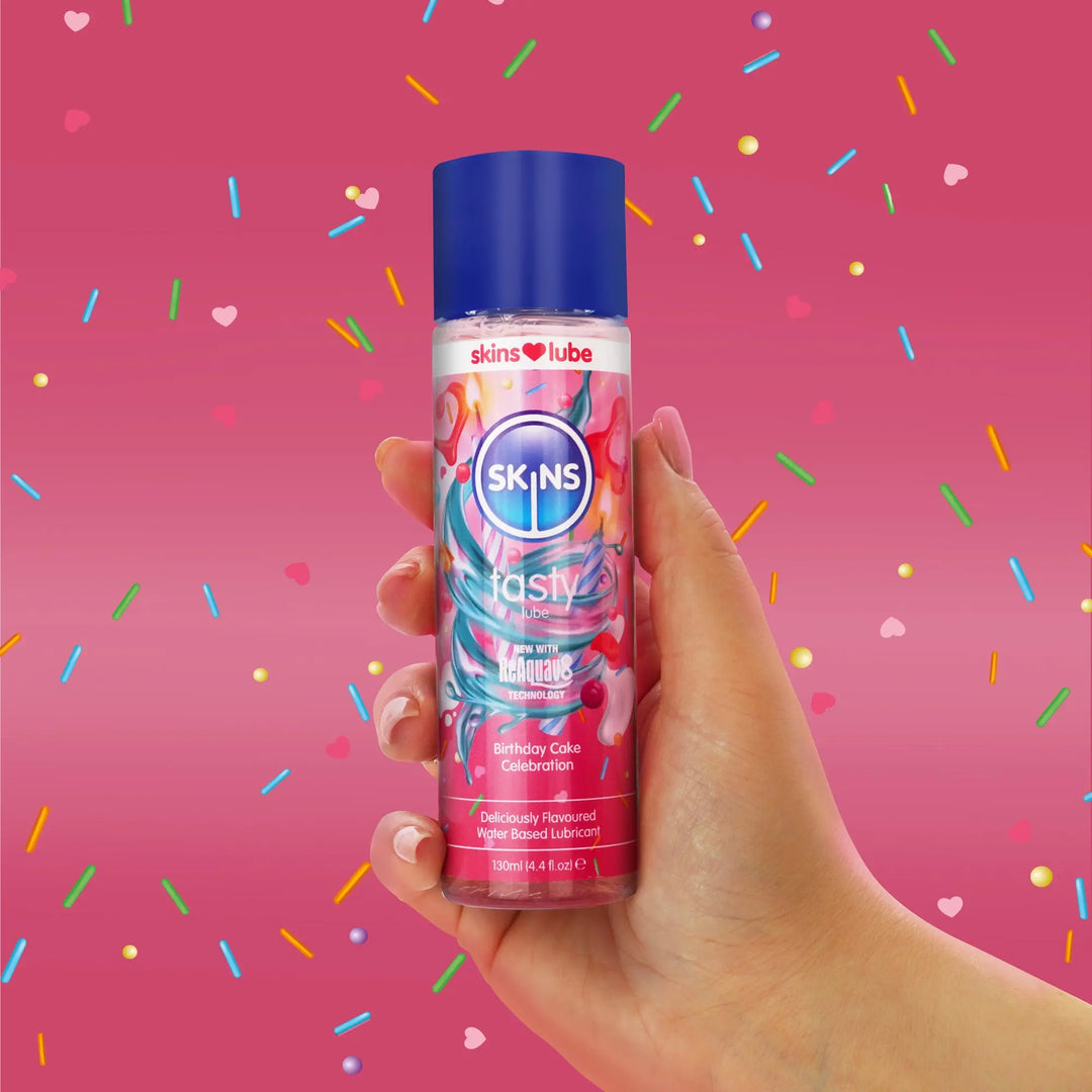 Hand holding a Skins birthday cake lubricant bottle against a pink background with colourful sprinkles.